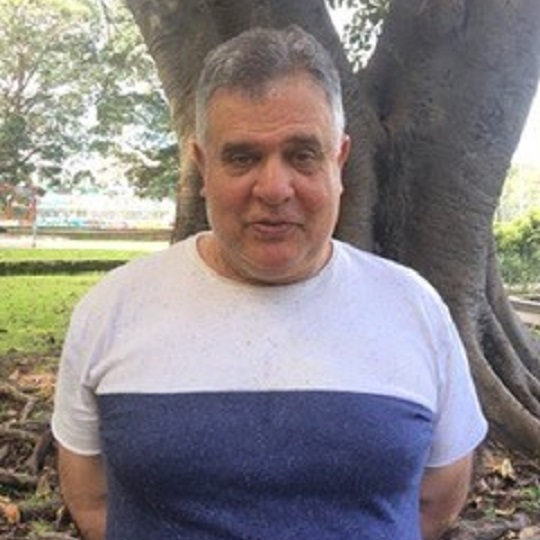 Photo of artist Vonwiller in park. Photo is head and shoulder of Paul wearing a white tshirt with navy blue panel. He is in front of a large morton bay fig tree